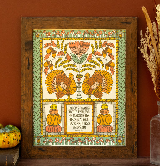 The O Give Thanks art print features the hand-lettered text of Psalm 107:1 surrounded by autumnal turkeys and squashes; shown in a frame and styled with a vase of dried grasses and a pumpkin figurine.