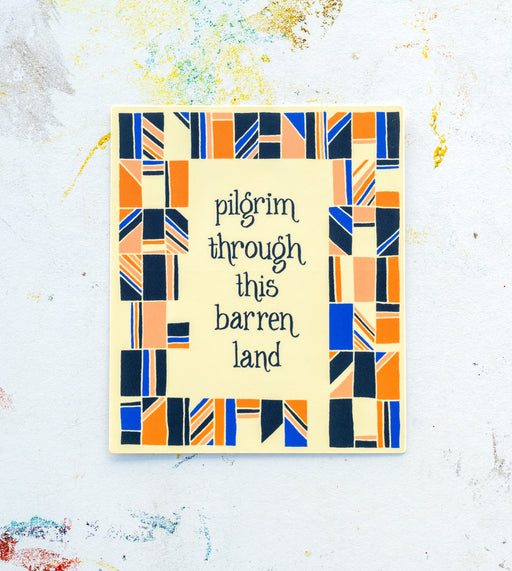 The “Pilgrim through this Barren Land” hymn sticker features hand lettered text surrounded by whimsical geometrical design in hues of blue, pink, black, and cream; displayed here on a white worktable background.