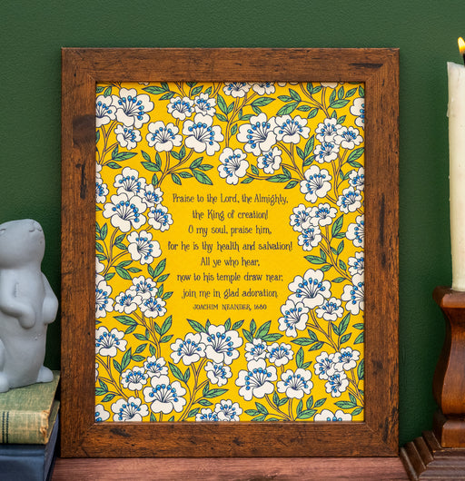 The "Praise to the Lord, the Almighty" hymn art print features hymn text surrounded by white and blue florals against a buttercup yellow. Shown here framed and styled with a ceramic rabbit, books, and a wooden candle holder.