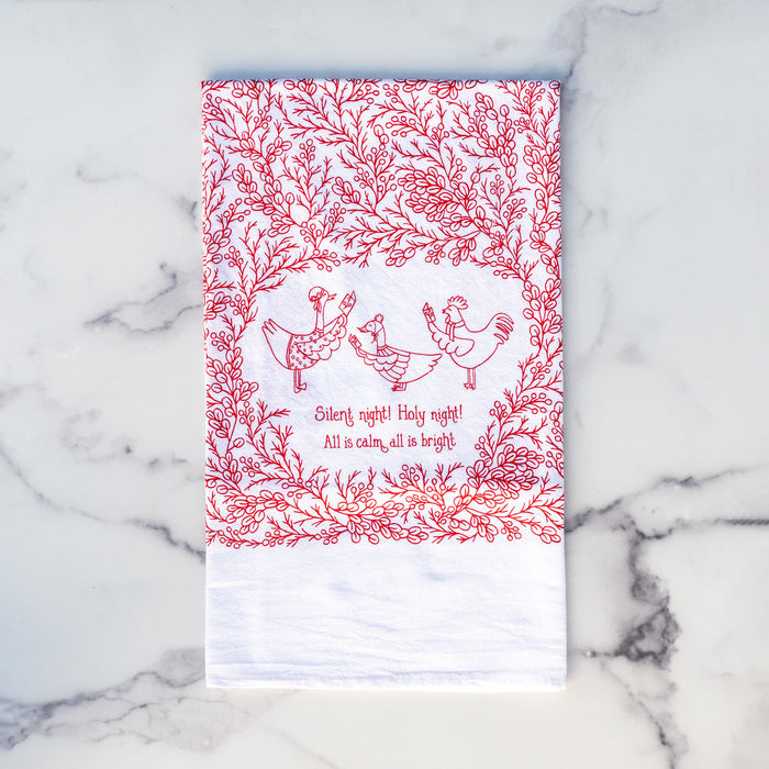 Our Silent Night Christmas hymn tea towel features a hand-illustrated floral border and hand-lettered text, printed in holly berry red on a 100% cotton tea towel, shown folded against a marble background