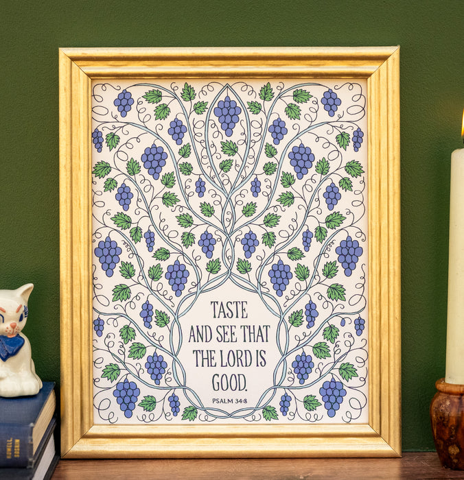 The Taste and See that the Lord Is Good art print features hand-lettered text from Psalm 34:8 surrounded by sprawling vines and grapes; shown framed and styled with books, a figurine, and a candle.