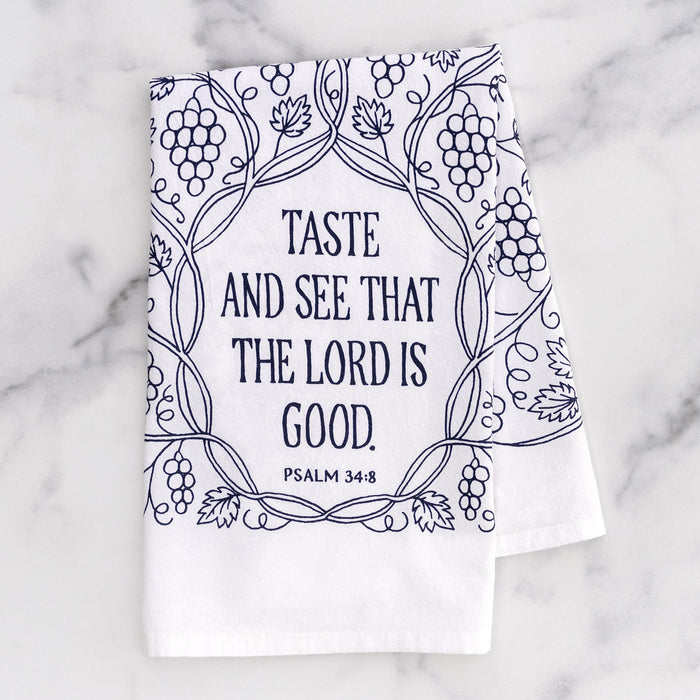 The Taste and See that the Lord Is Good Scripture tea towel is printed in concord purple and features Psalm 34:8; it is shown here folded against a gray marble background.