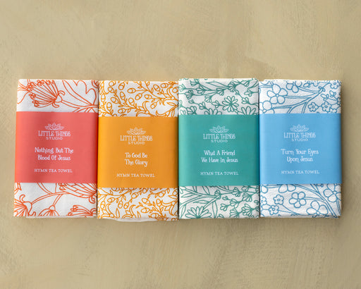 Set 1 of Little Things Studio hymn tea towels featuring four classic hymns with floral designs printed on a 100% cotton tea towels, shown folded and wrapped with paper belly bands set against a khaki background.