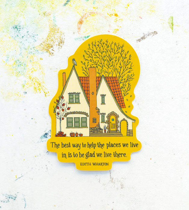 "The Best Way to Help” sticker features the hand-lettered quote by Edith Wharton coupled with a delightfully autumnal house illustration and a sunset orange background, shown against the backdrop of a white worktable.