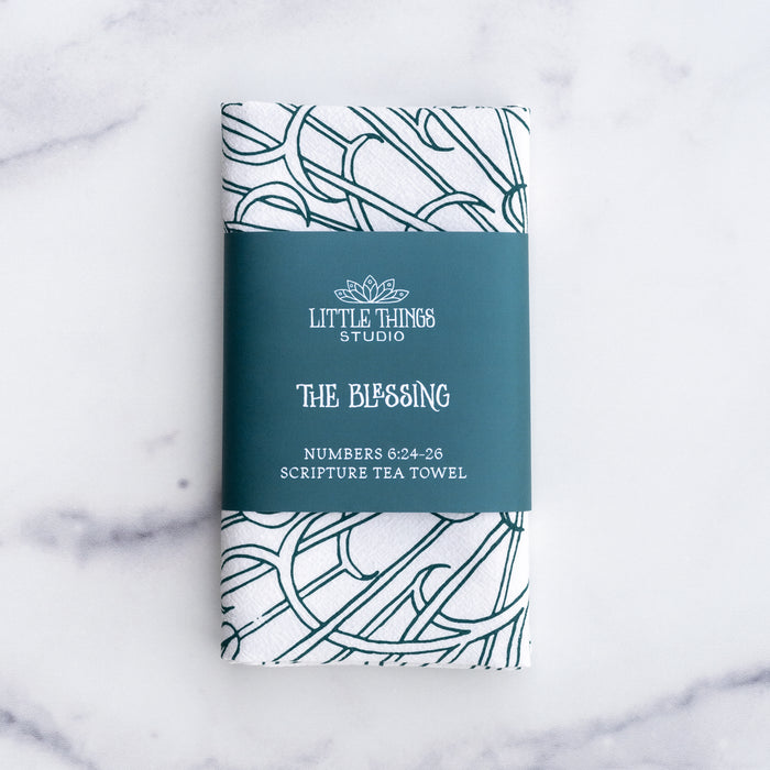 The Blessing Scripture tea towel is printed in muted blue-green tone and features the verse from Numbers 6:24–26. Made of 100% cotton. Shown folded and wrapped with a paper belly band for gift giving.