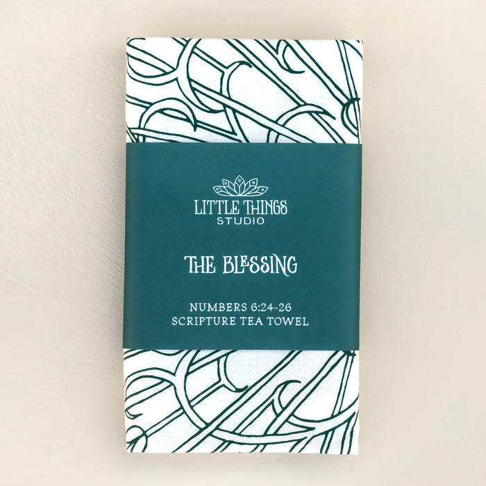 The Blessing Scripture tea towel is printed in muted blue-green tone and features the verse from Numbers 6:24–26. Made of 100% cotton. Shown here folded with a paper belly band for gift giving, against a khaki background.