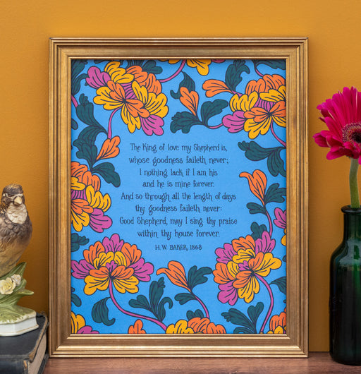 The King of Love My Shepherd Is art print features the cherished hymn surrounded by vibrant jewel-toned floral illustrations against a bright blue background; shown here in a frame and styled with an owl figurine, books, and a flower in a vase.