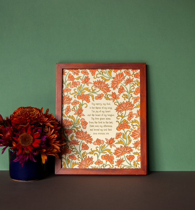 Thy Mercy My God art print, featuring hand-lettered text on a cream background with hand illustrated floral detail in shades of orange, pink, gold, and green, is displayed in a dark wood frame next to a vase of fresh flowers.