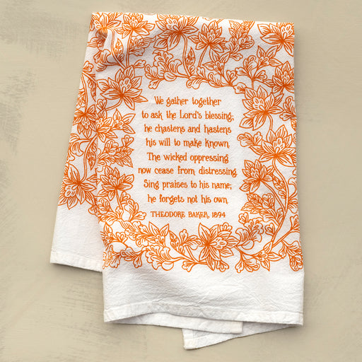 The We Gather Together hymn tea towel is printed in autumn orange and features hand lettering and floral illustration on a 24" x 20", 100% cotton towel. Shown folded against a khaki background.