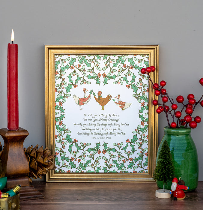 We Wish You a Merry Christmas carol art print features illustration of lovely holly branches with festive animal carolers against a cream background, displayed in a burnished gold frame, styled alongside holly berries in a vase with a festive candle, pinecone, and other figurines.