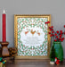 We Wish You a Merry Christmas carol art print features illustration of lovely holly branches with festive animal carolers against a cream background, displayed in a burnished gold frame, styled alongside holly berries in a vase with a festive candle, pinecone, and other figurines.