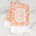 The We Gather Together hymn tea towel is printed in autumn orange and features hand lettering and floral illustration on a 24" x 20", 100% cotton towel. Shown folded against a white marble background.