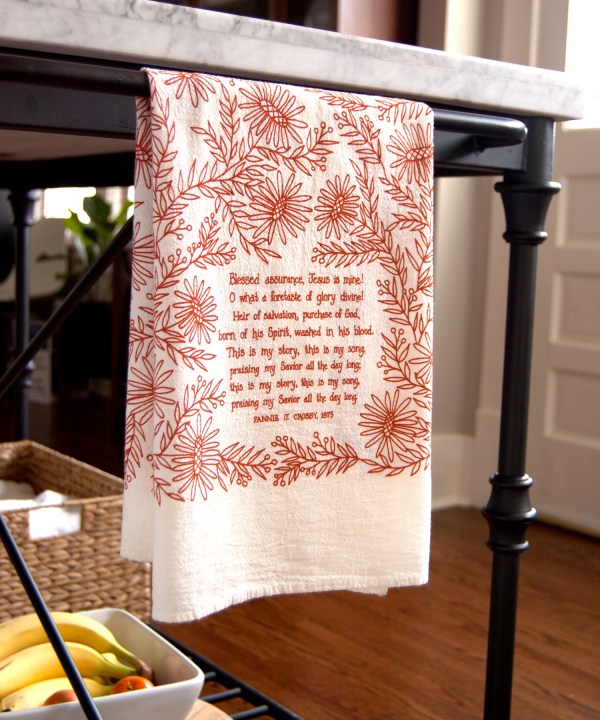 The Blessed Assurance tea towel is printed in rusty red and features hand lettered hymn text surrounded by intricate floral illustration. It is pictured here folded and hanging on a kitchen towel rack