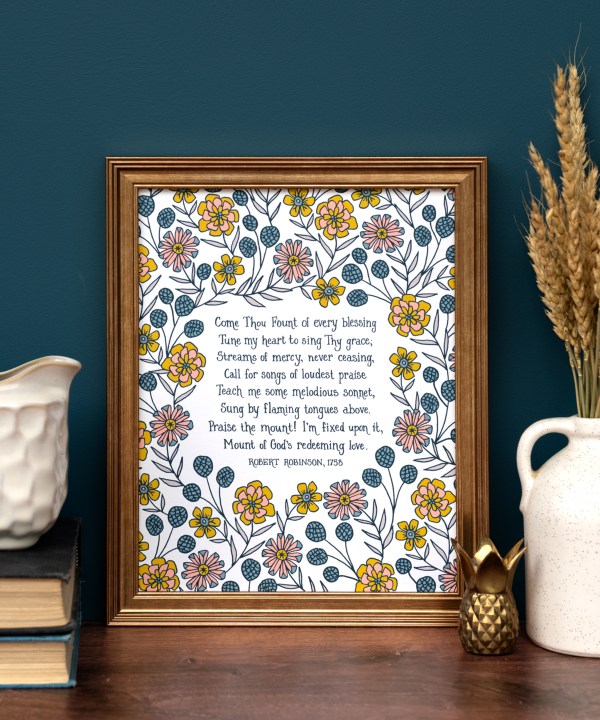 Come Thou Fount hymn art print — faith based wall decor featuring hand lettered text on a white background accented by illustrated floral design in yellow, orange, teal, and green, displayed in a wood frame with a vase of dried grasses, stack of books and a ceramic creamer.