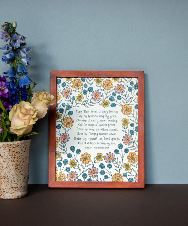 Come Thou Fount hymn art print — faith based wall decor featuring hand lettered text on a white background accented by illustrated floral design in yellow, orange, teal, and green, displayed in a dark wood frame with a vase of fresh flowers