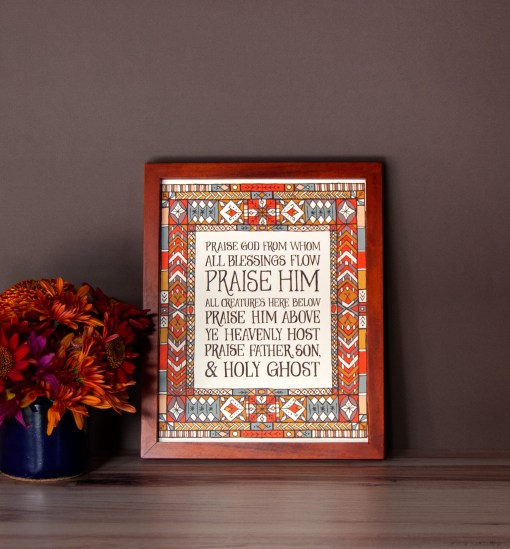 Doxology wall art — a Christian picture for wall featuring hand lettered text surrounded by a multi-coloured stained glass illustration, displayed in a dark wood frame with a vase of fresh flowers