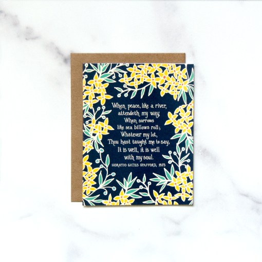 The It is Well with My Soul greeting card features bold yellow floral border surrounding the hymn text against a navy blue background; pictured here against a white marble backdrop.