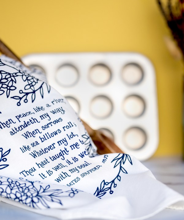 It Is Well With My Soul tea towel, printed in a striking cobalt blue and is shown folded and styled with a wooden spoon, vase with dried flowers, and muffin tin.