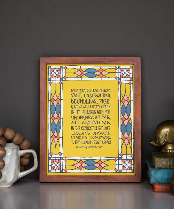 o the deep deep love of jesus wall art styled with stained glass border in blue, pink, red and cream, printed on a mustard background, displayed in a dark wood frame with books, figurine and mug