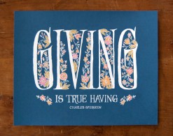 Giving is True Having quote print — Charles Spurgeon quote artistically displayed with hand lettered text, printed on a navy background and accented by delicate floral illustration in shades of pink, orange, and cream, displayed against a dark wood backdrop