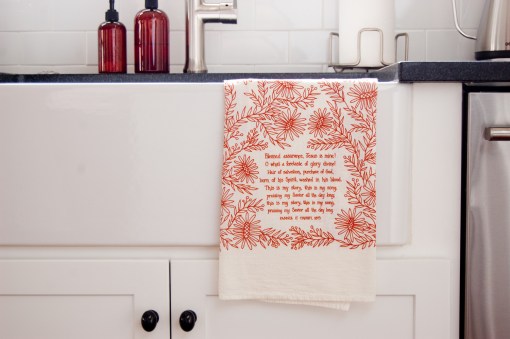 The Blessed Assurance tea towel is printed in rusty red and features hand lettered hymn text surrounded by intricate floral illustration. It is pictured here folded and hanging on a kitchen sing
