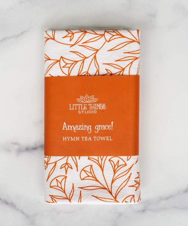 Amazing Grace tea towel is one of our hymn tea towels, printed in pumpkin orange and featuring hand illustrated floral designs surrounding the hand lettered text, shown folded and displayed wrapped with a paper belly band and ready for gift giving.