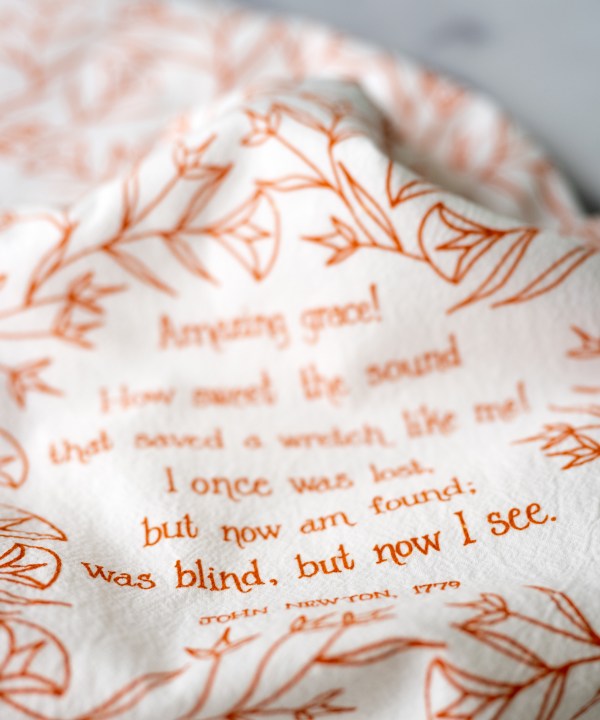 Floral and text detail of the Amazing Grace tea towel is one of our hymn tea towels, printed in pumpkin orange and featuring hand illustrated floral designs surrounding the hand lettered text.