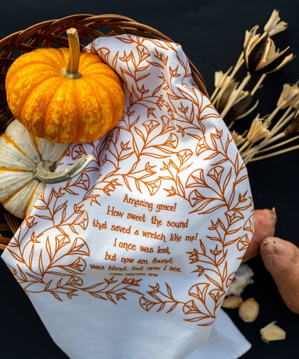 Amazing Grace tea towel is one of our hymn tea towels, printed in pumpkin orange and featuring hand illustrated floral designs surrounding the hand lettered text, shown styled with mini pumpkins and dried grasses.