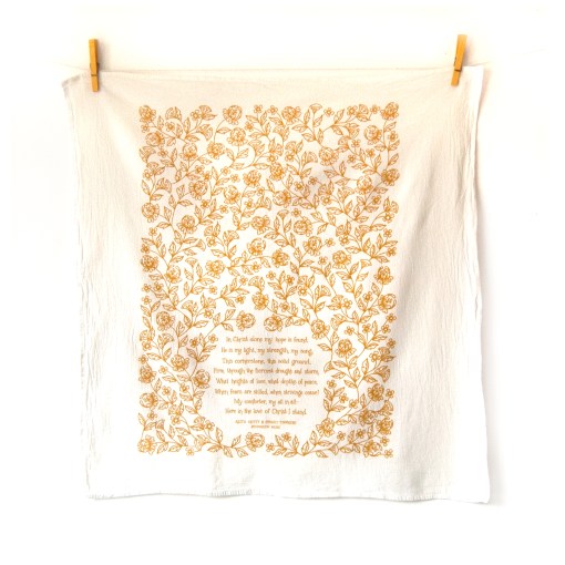 The In Christ Alone tea towel is printed in honey yellow and features floral illustrations surrounding the hand-lettered hymn text. Pictured unfolded and hanging with clothes pins.