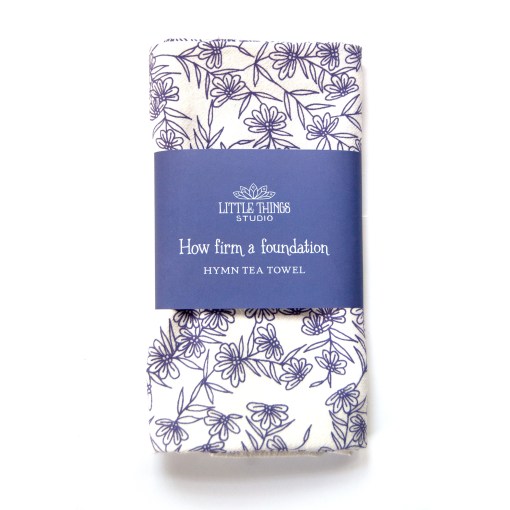 How Firm a Foundation hymn tea towel is printed in periwinkle and displayed folded with a printed paper belly band for gift giving