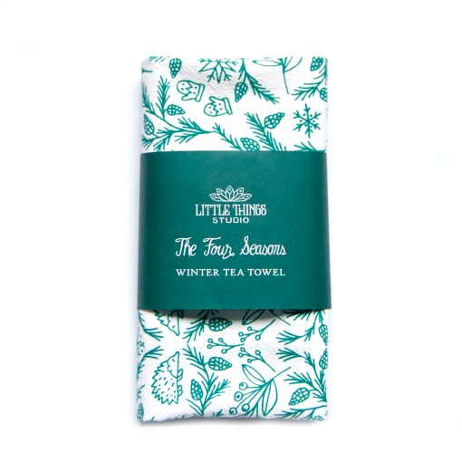 The winter tea towel is printed in pine needle green and features whimsical seasonal illustrations surrounding a winter cottage. Shown folded and wrapped with a paper belly band for gift giving.