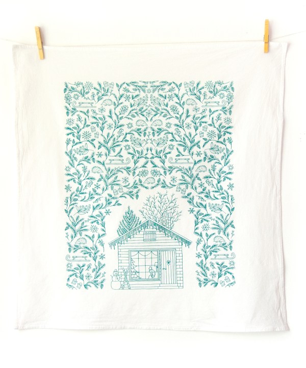 The winter tea towel is printed in pine needle green and features whimsical seasonal illustrations surrounding a winter cottage — could be used as a Christmas tea towel. Shown here unfolded and hanging with clothes pins.