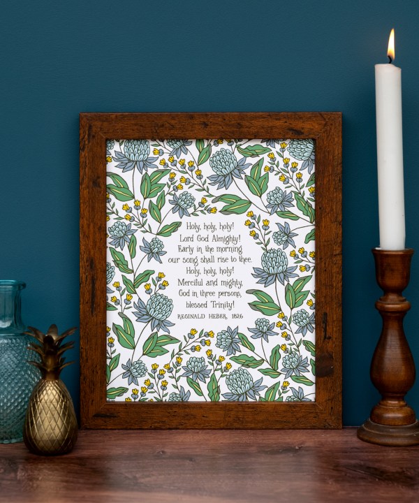 Holy Holy Holy art print — biblical wall art featuring hand lettered text surrounded by floral illustrations in shades of blue, yellow, and green, displayed in a dark wood frame with a wooden candlestick and some vases