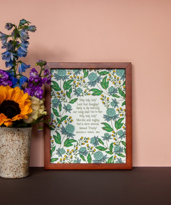 Holy Holy Holy art print — biblical wall art featuring hand lettered text surrounded by floral illustrations in shades of blue, yellow, and green, displayed in a dark wood frame with a vase of vibrant fresh flowers