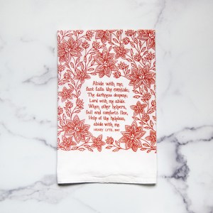 Abide with Me hymn tea towel printed in apple red, pictured folded against a white marble background