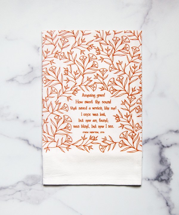 Amazing Grace tea towel is one of our hymn tea towels, printed in pumpkin orange and featuring hand illustrated floral designs surrounding the hand lettered text, shown folded and displayed against a white marble background.