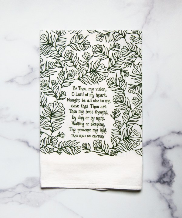 The Be Thou My Vision tea towel is printed in forest green and features hand lettered text surrounded by illustrated botanical detail, displayed folded against a white marble background.
