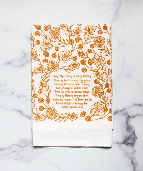 Come Thou Fount hymn tea towel is printed in butterscotch and features hand lettered hymn text surrounded by illustrated florals, pictured folded against a white marble background.
