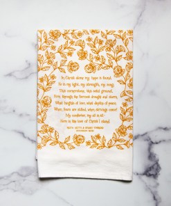 The In Christ Alone tea towel is printed in honey yellow and features floral illustrations surrounding the hand-lettered hymn text. Pictured folded against a white marble background.