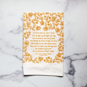 The In Christ Alone tea towel is printed in honey yellow and features floral illustrations surrounding the hand-lettered hymn text. Pictured folded against a white marble background.