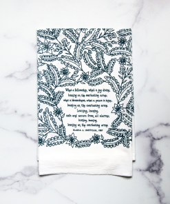 Leaning on the Everlasting Arms tea towel is printed in dark teal and features hand lettered hymn text surrounded by illustrated florals, pictured here folded against a white marble background.
