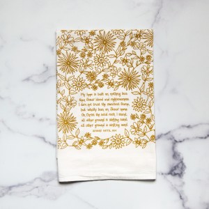 The Hope is Built on Nothing Less tea towel is printed in burnished gold and features the hand lettered hymn text surrounded by intricate floral illustrations, pictured folded against a white marble background