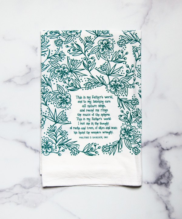 This is My Father's World tea towel is printed in teal and features hand lettered hymn text surrounded by intricate floral illustration, pictured folded against a white marble background