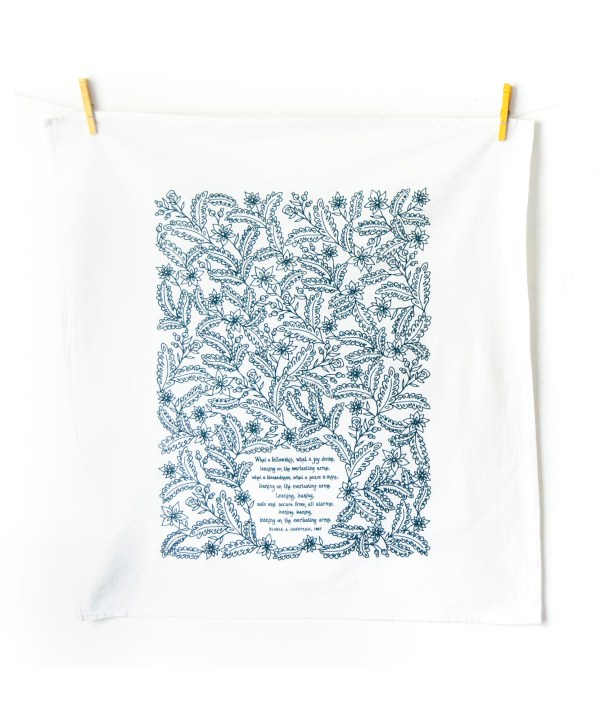 Leaning on the Everlasting Arms tea towel is printed in deep sea blue and features hand lettered hymn text surrounded by illustrated florals, pictured here unfolded and hanging with clothes pins against a white background.