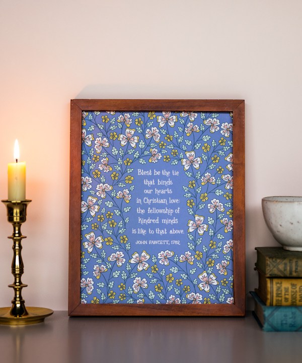 Blest be the Tie hymn art print, featuring hand lettered text offset by delicate hand illustrated floral designs, printed on a periwinkle blue background, displayed in a dark wood frame with a candle and stack of books