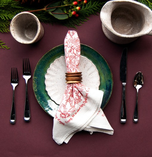 "O Come All Ye Faithful" cloth napkins use the same flour sack cotton as our tea towels and are printed in a vibrant Christmas red with a beautiful floral illustration and the text printed around the edge. Shown styled with a napkin ring and festive place setting.