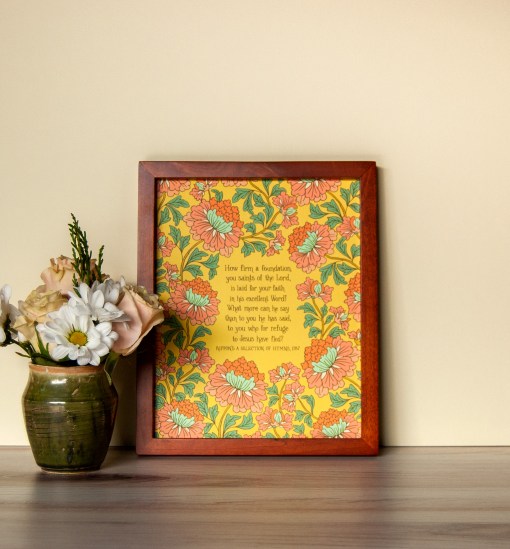 How Firm a Foundation art print — hand-lettered hymn text on a mustard background surrounded by a floral design using light red, light blue and green, pictured in a dark wood frame next to a vase of fresh flowers