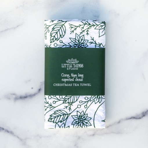 Come Thou Long Expected Jesus Christmas/advent tea towel is hand-lettered with a floral border and printed in pine green on a 100% cotton tea towel, shown wrapped with a paper belly band for gift giving.