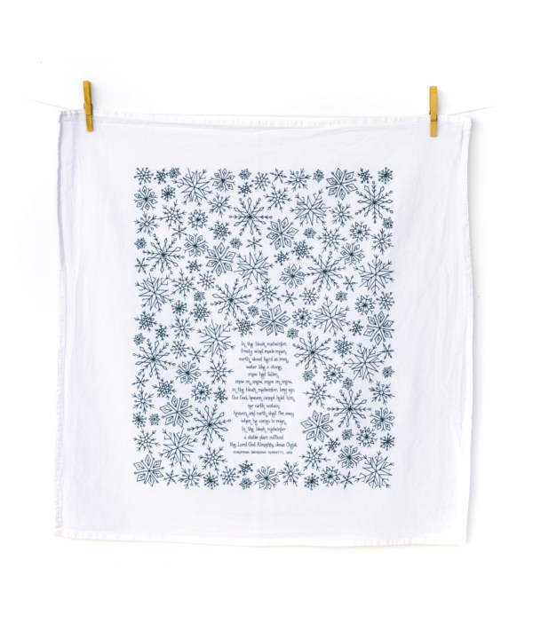 In the Bleak Midwinter Christmas tea towel is hand-lettered with a floral border and printed in evergreen on a 100% cotton tea towel, shown hanging with clothes pins.