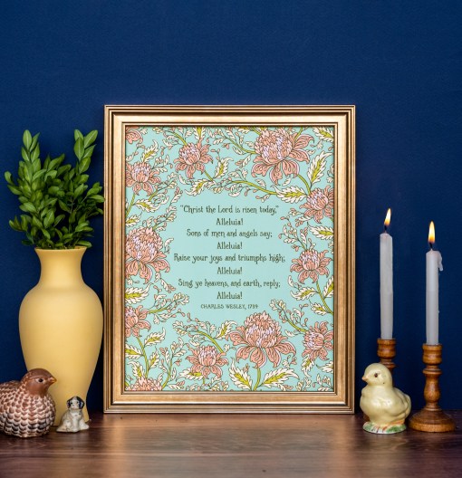 Hymn art print of Christ the Lord is Risen today, featuring hand-lettered hymn text and hand illustrated florals in corals and pinks against a mint green background, displayed in a gold frame and styled with vase of fresh greens, candles, and ceramic figurines against a navy wall.